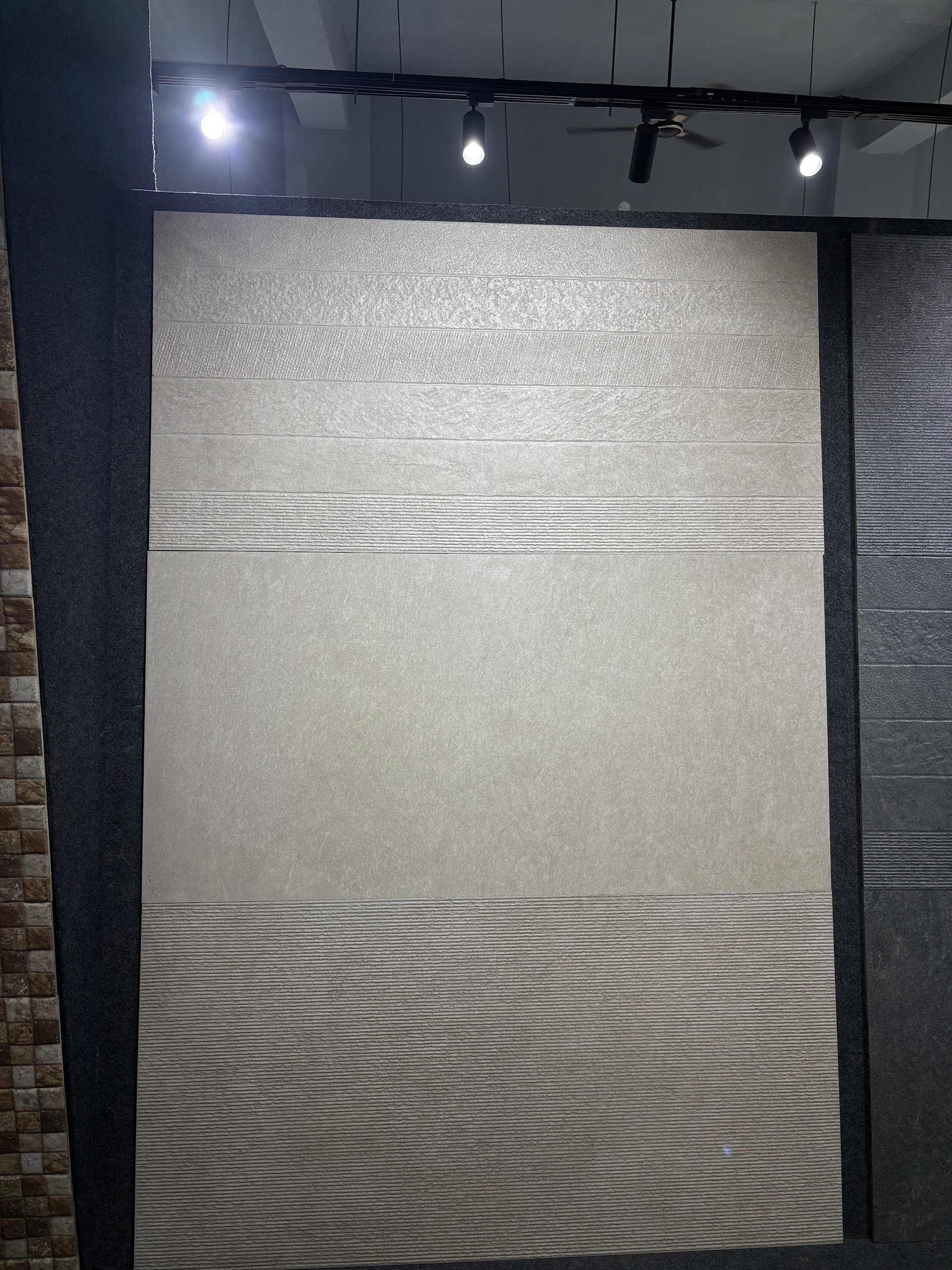 Pale neutral tile display with fine texture and a contrasting lower band under showroom spotlights