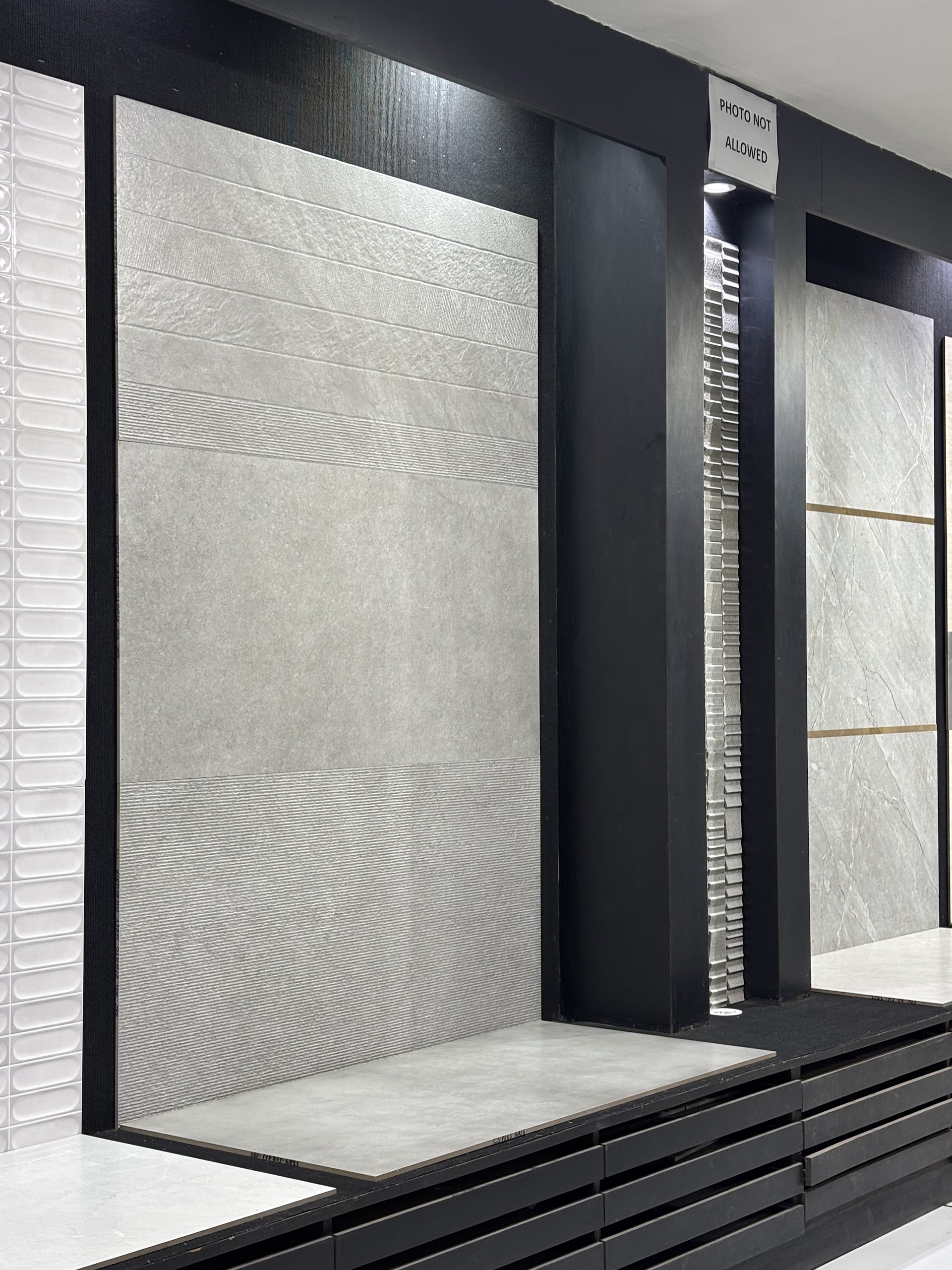Pale textured wall tile panel with a matching horizontal display sample in a black-framed showroom bay