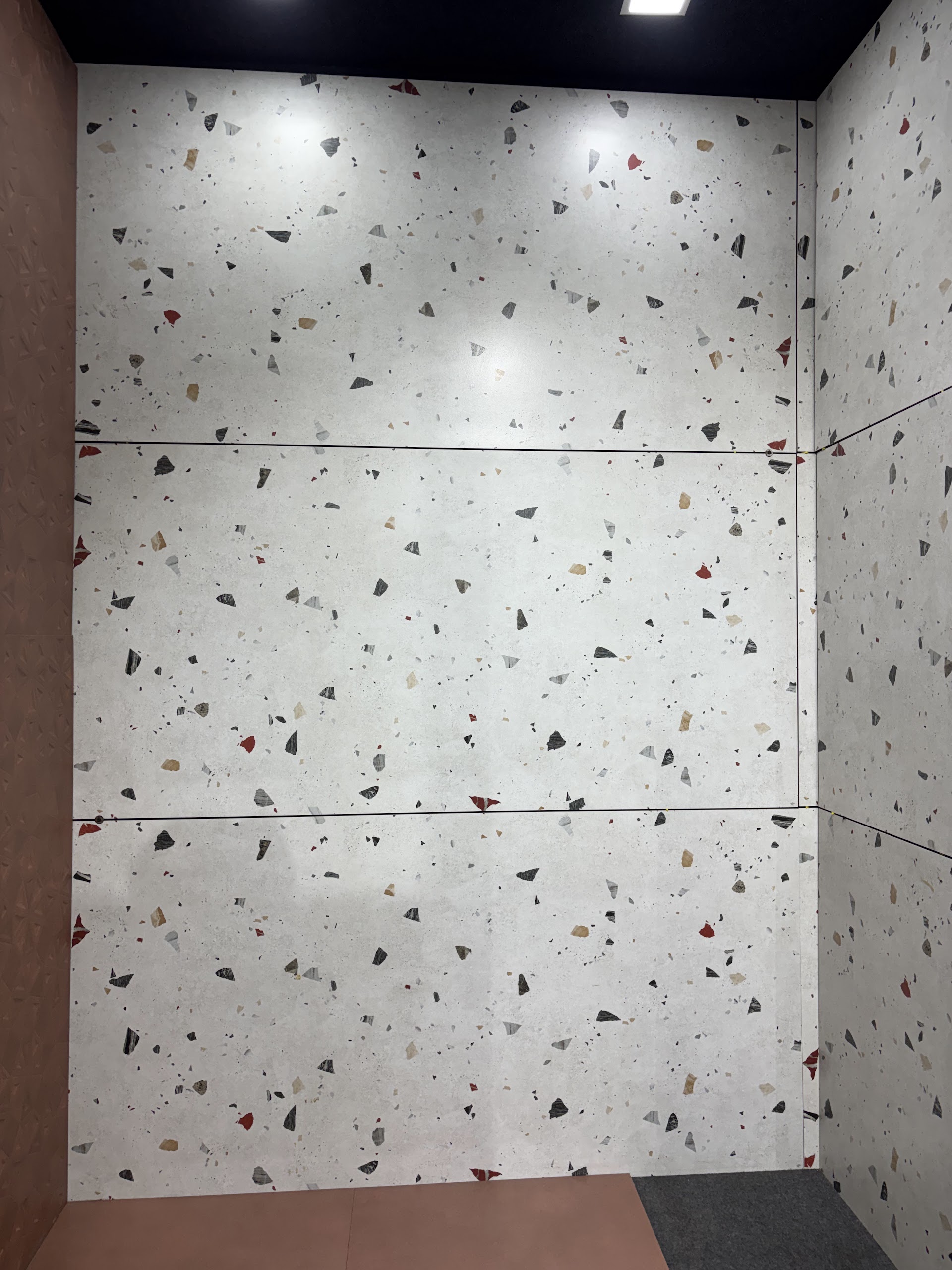 White terrazzo-finish tile wall with scattered dark and muted coloured fragments across several joined panels